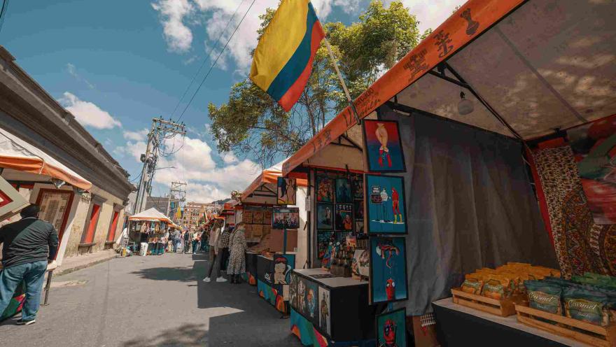 Calle con toldos de comercio en los que hay artesanías, cuadros y personas observando en algunos de ellos. Destaca una bandera de Colombia colgada en uno de los locales