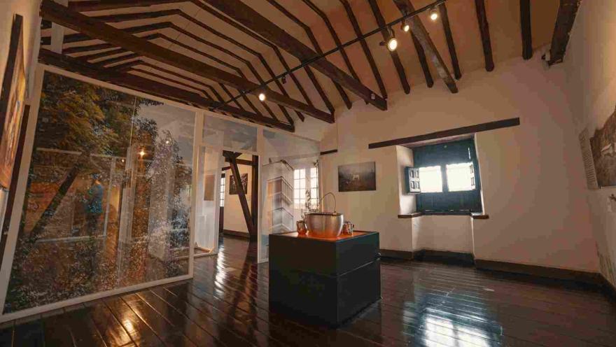 Un salón interior en el que sobresale el techo en vigas de madera y, en el centro del piso, sobre una plataforma, una olla sopera grande. Al fondo hay una pequeña ventana por donde entra algo de luz natural.