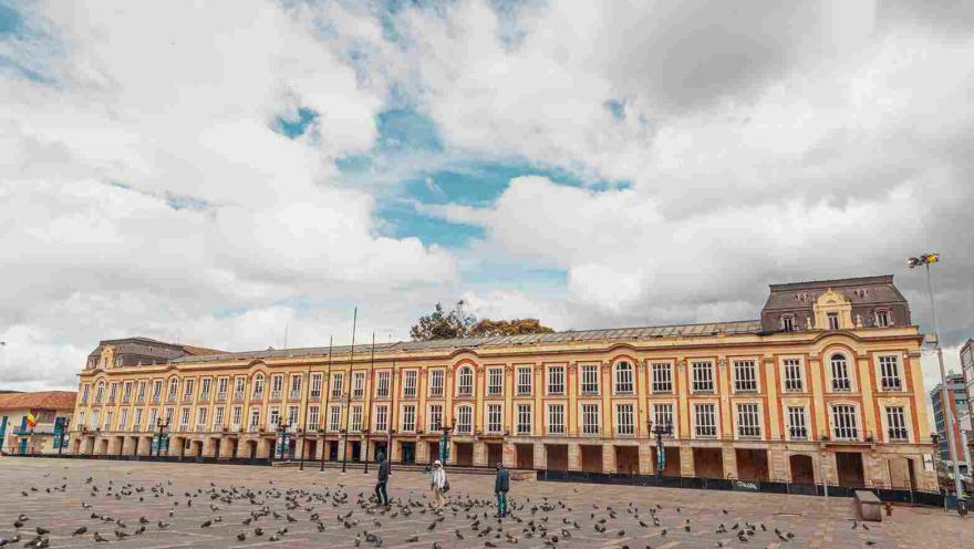 Plano general de la fachada exterior del palacio, vista de frente. Se observan muchas puertas en piedra en el primer piso y muchas ventanas en el segundo. Al frente se ve la Plaza de Bolívar con algunas personas y varias palomas.