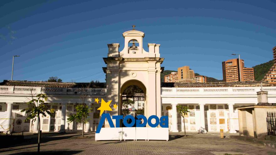 Entrada principal del Cementerio Central en la que destaca una alta puerta en forma de arco, con un campanario y reloj. Al frente hay una gran estructura con la palabra 'Bogotá' en letras azules y con una estrella amarilla encima de la letra A.