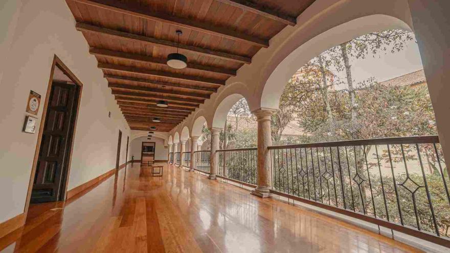 Un pasillo largo en un segundo piso en donde resalta el piso en madera brillante y varias columnas que forman arcos que sostienen el techo. Al fondo se observa mucha vegetación.