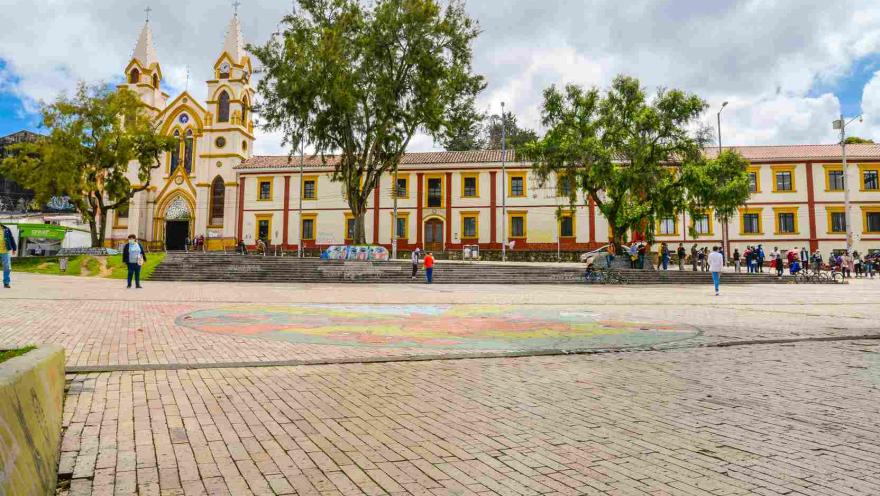 Fachada de iglesia grande de color curuba y franjas rojas. Al frente hay una plazoleta con algunas personas que parece disfrutan del lugar.