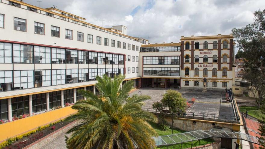 Fachada del Museo de La Salle, un conjunto arquitectónico que combina un edificio histórico de ladrillo y arcos con una estructura moderna de amplias ventanas. En el centro se abre un patio amplio y despejado, rodeado de zonas verdes y una palma que aporta frescura al entorno. El cielo parcialmente nublado enmarca el espacio, resaltando el carácter académico y cultural del lugar.