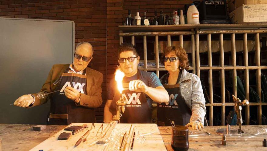 Tres personas, dos hombres y una mujer, trabajan con piezas de vidrio. El hombre en el centro calienta y da forma a un vidrio con fuego.