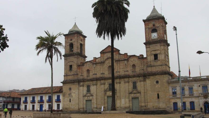 Facade of the Cathedral of the Holy Trinity and Saint Anthony of Padua in Zipaquirá. The structure is tall, made of stone, and has two towers at either end. The clock is in the left tower, and the bell tower is in the right. In front are two tall palm trees and a small plaza.