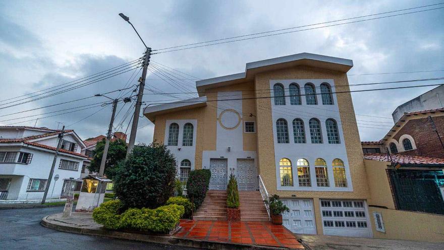 Fachada de una casa grande de estilo residencial con acabados en tonos beige y blanco, ubicada en una esquina de barrio. El edificio tiene varias ventanas arqueadas, una escalinata central que conduce a la entrada y un garaje en la parte inferior.