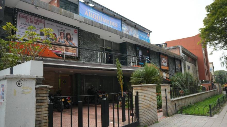 Facade of a cultural building or theater in an urban area, with advertising posters for plays at the top. The building has two levels, brick and stone walls, and an entrance with a black metal gate. A small garden with ornamental plants and a tree-lined walkway are visible in front, to the right.