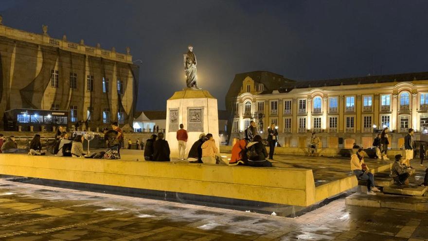 Plaza pública en horario nocturno con una estatua iluminada en el centro, rodeada de personas sentadas y conversando sobre una plataforma. Los edificios históricos alrededor están iluminados, destacando su arquitectura colonial. El suelo húmedo refleja las luces cálidas, creando un ambiente urbano animado y acogedor durante la noche.