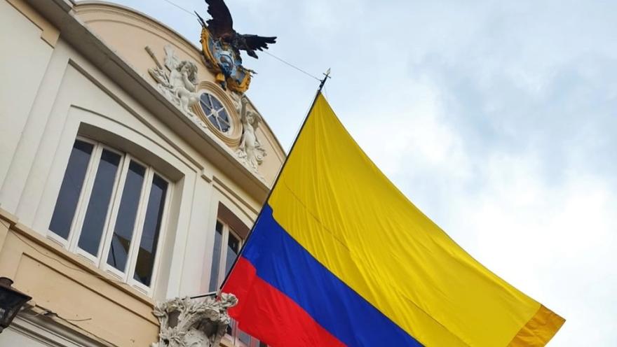  Fachada de un edificio histórico con relieves decorativos y una escultura de un condor en la parte superior, junto a una gran bandera de Colombia ondeando frente al cielo.