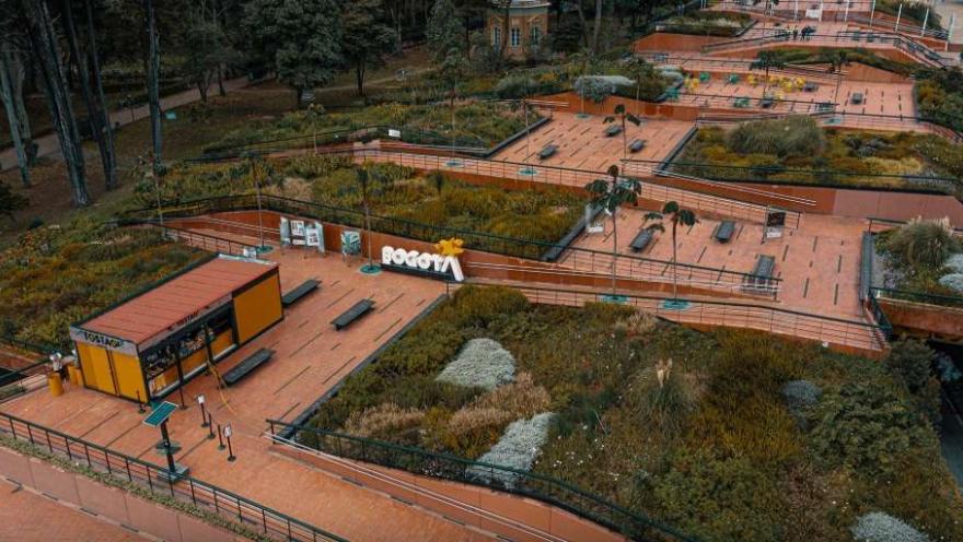 Vista aérea de un parque urbano con senderos amplios en tonos rojizos, rodeados de jardines y vegetación variada. Se observan bancas, barandas y terrazas escalonadas, además de un pequeño módulo o quiosco y un letrero que dice “Bogotá”. El entorno es verde y organizado, con áreas para el descanso y el recorrido peatonal.