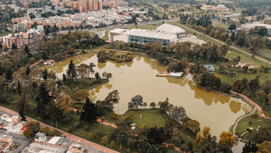 Vista aérea de un parque urbano con un gran lago rodeado de árboles, senderos peatonales y zonas verdes, con edificios y barrios de la ciudad alrededor.