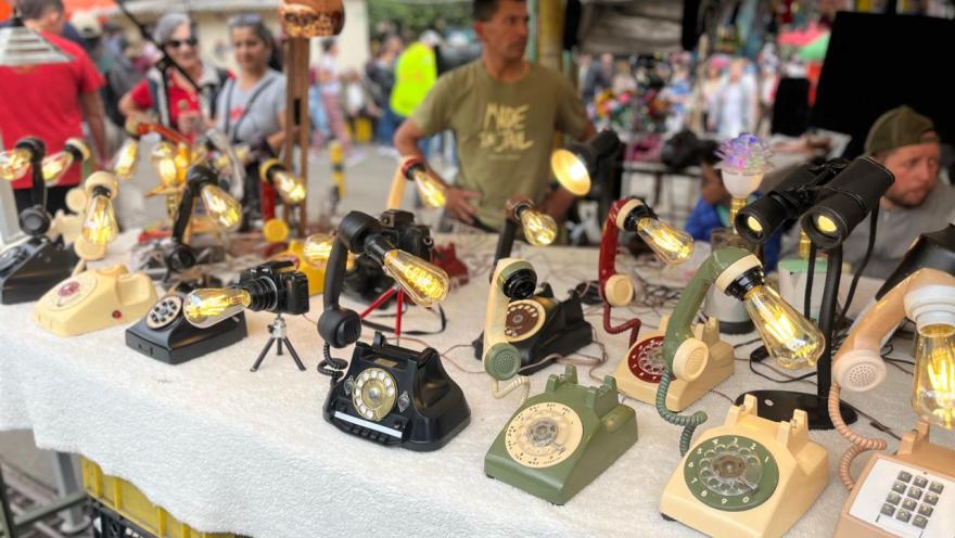 An open-air market stall with a covered table displaying vintage rotary dial telephones converted into lamps, with lit bulbs. The objects are arranged in rows and showcase various vintage colors and designs.