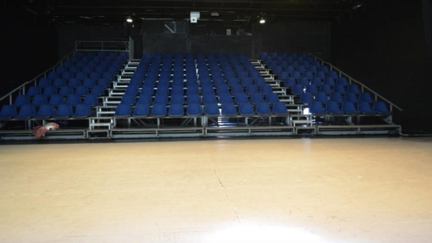  Interior de una sala de teatro con graderías metálicas y filas de sillas azules dispuestas en forma escalonada. El escenario se observa en primer plano, vacío y bien iluminado, mientras el fondo es oscuro, con iluminación técnica en el techo. El espacio transmite un ambiente preparado para funciones escénicas.