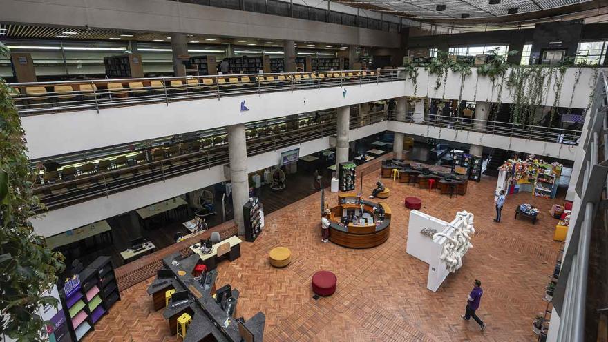 Vista interior de una biblioteca amplia de varios niveles, con balcones que rodean un gran espacio central. En el primer piso hay mesas, estanterías, zonas de lectura y algunos visitantes dispersos. El suelo es de ladrillo en patrón geométrico y en los niveles superiores se observan filas de libros y puestos de estudio.