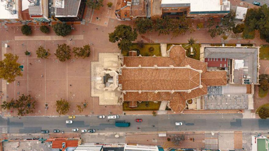 Vista aérea cenital de una iglesia con techo de tejas rojas, rodeada por una amplia plaza peatonal con árboles dispersos. A un lado se observa una vía con tráfico de carros y, alrededor, edificaciones urbanas. La composición muestra la organización del espacio público y la arquitectura del sector desde una perspectiva superior.