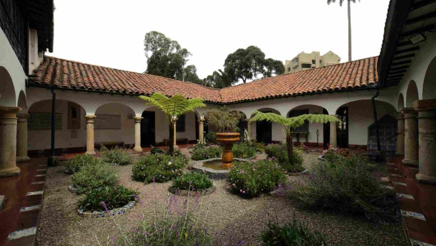 Patio interior de arquitectura colonial con corredores de arcos y columnas, techos de teja roja y un jardín central con plantas, helechos y una pequeña fuente de piedra en el centro.