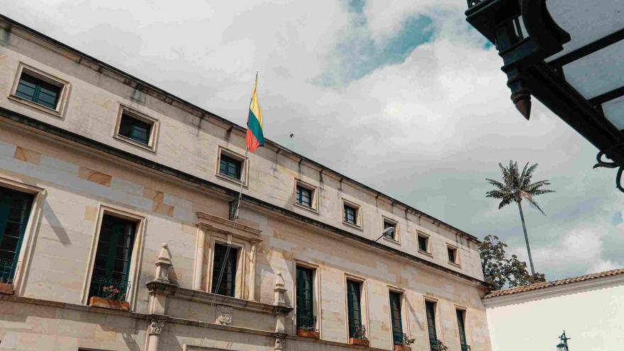 Fachada de un edificio histórico de estilo colonial con muros en tonos claros y ventanas alineadas con marcos oscuros y balcones con flores. En el centro ondea la bandera de Colombia sobre la entrada principal. El cielo está parcialmente nublado y a un costado se eleva una palma, mientras un elemento arquitectónico decorativo aparece en primer plano.