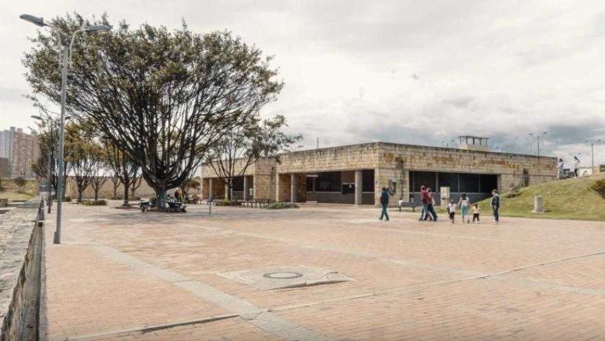 Plaza amplia en ladrillo al aire libre, con un edificio bajo de piedra y ventanales oscuros al fondo. A la izquierda, un árbol grande brinda sombra junto a bancas y faroles, mientras varias personas caminan por el espacio abierto en un entorno urbano con cielo nublado.