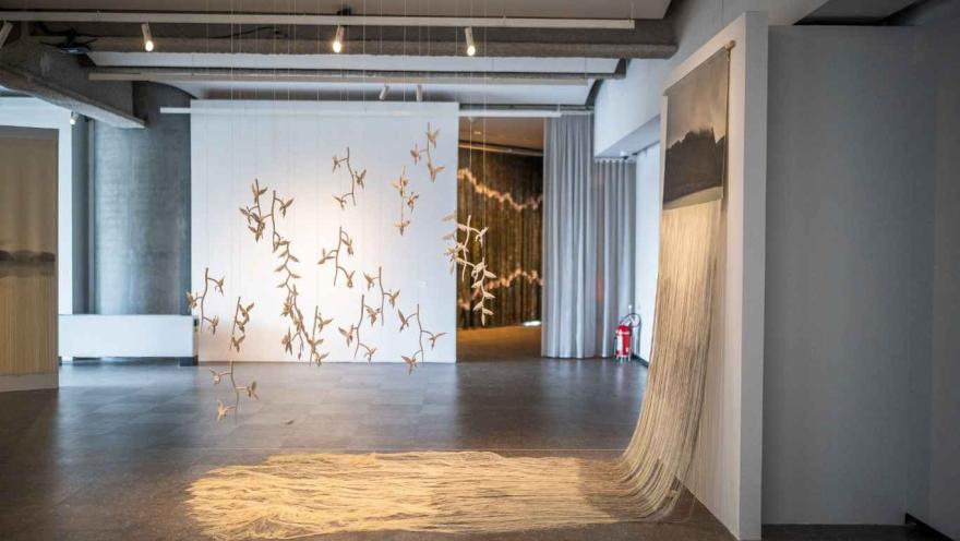 Instalación artística contemporánea en la Sala de Arte Bancolombia, donde delicadas figuras suspendidas cuelgan del techo frente a un muro blanco, mientras hilos o fibras textiles caen desde una estructura formando una especie de manto sobre el suelo.