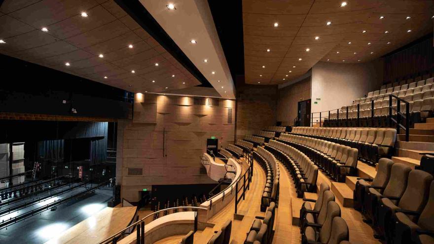Interior de un teatro moderno con graderías semicirculares de asientos acolchados que miran hacia un escenario iluminado. El recinto tiene paredes en tonos cálidos, barandas negras y un techo con múltiples luces empotradas que iluminan el espacio.