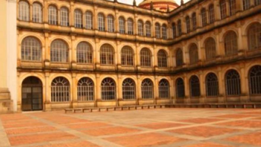  Patio amplio de un edificio histórico con arquitectura colonial, rodeado por una estructura de varios pisos con arcos repetidos y grandes ventanales. El suelo es de ladrillo dispuesto en patrón geométrico y el espacio está casi vacío, con una atmósfera tranquila. 