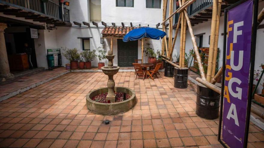 Patio interior de una casa colonial con piso de baldosas de barro, una fuente de piedra en el centro y mesas de madera con sombrilla azul. Alrededor hay balcones con barandas negras, plantas en macetas y una estructura de guadua junto a un cartel morado que dice “FUGA”.