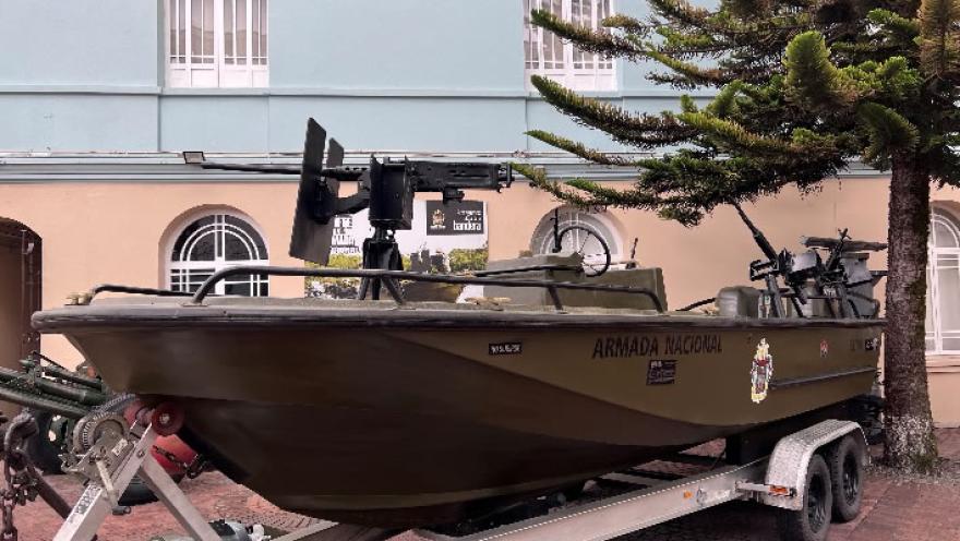 Embarcación militar de la Armada Nacional montada sobre un remolque, equipada con ametralladoras y exhibida al aire libre frente a un edificio.