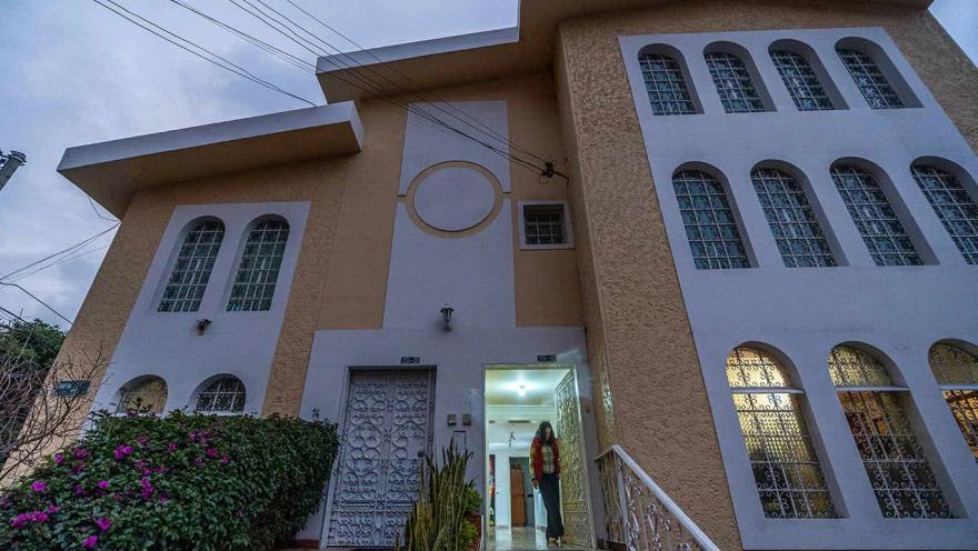Vista cercana de la fachada de una casa de dos niveles con diseño en tonos beige y blanco, caracterizada por ventanas altas con arcos y rejas metálicas. En el centro hay una escalinata que conduce a la entrada principal, donde una persona se encuentra en la puerta iluminada. 