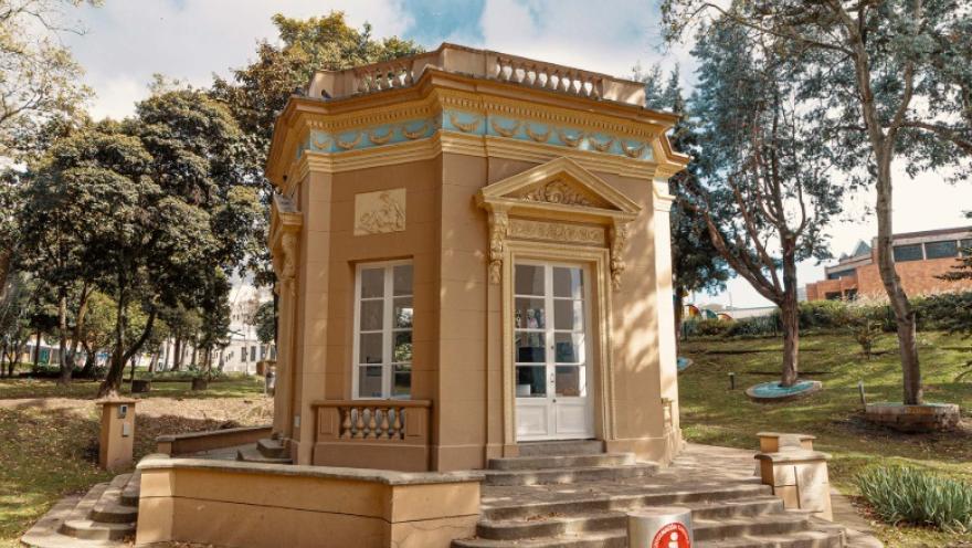 Pequeño quiosco de estilo clásico en medio de un parque, con fachada en tonos beige y detalles ornamentales en relieve y molduras decorativas en azul y dorado. El edificio tiene ventanas y una puerta blanca, además de una escalinata de acceso frontal. Está rodeado de árboles, zonas verdes y senderos, en un entorno tranquilo y luminoso durante el día.