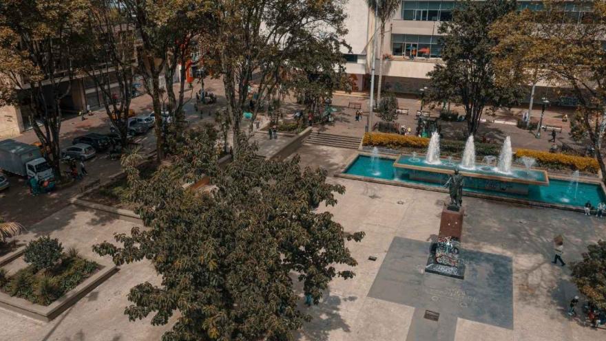Vista elevada de una plaza urbana con árboles frondosos, senderos peatonales y una fuente rectangular con chorros de agua en funcionamiento. En el centro se observa una estatua sobre pedestal, rodeada de espacios abiertos donde algunas personas caminan o descansan.