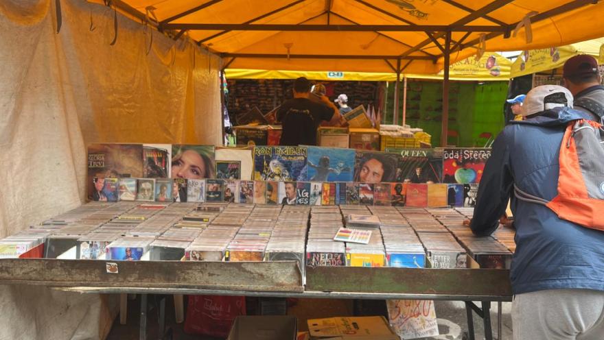 A market stall covered with a yellow tent displays records and CDs arranged in boxes on a table. Album covers from various artists are visible in the background, while some people browse the merchandise.