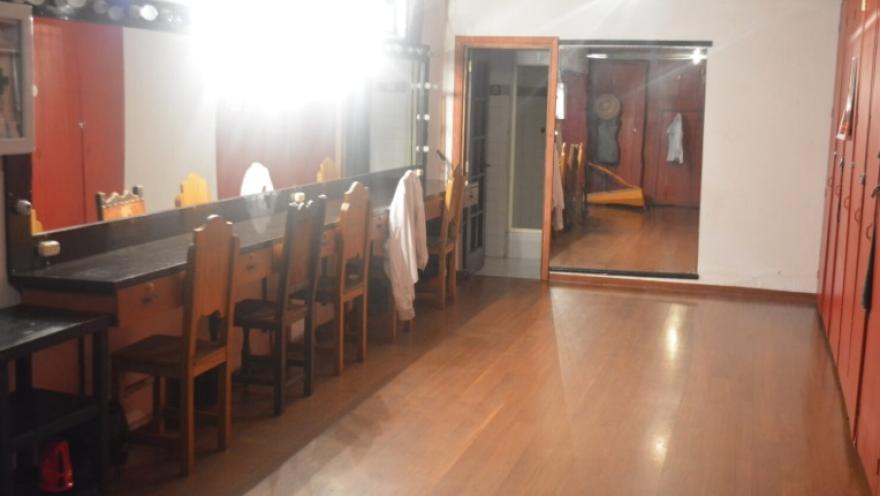 Interior de un camerino de teatro con piso de madera, equipado con un largo tocador con espejos iluminados y varias sillas de madera. A un lado se ven casilleros o armarios rojos, y al fondo un espejo grande refleja parte del espacio.