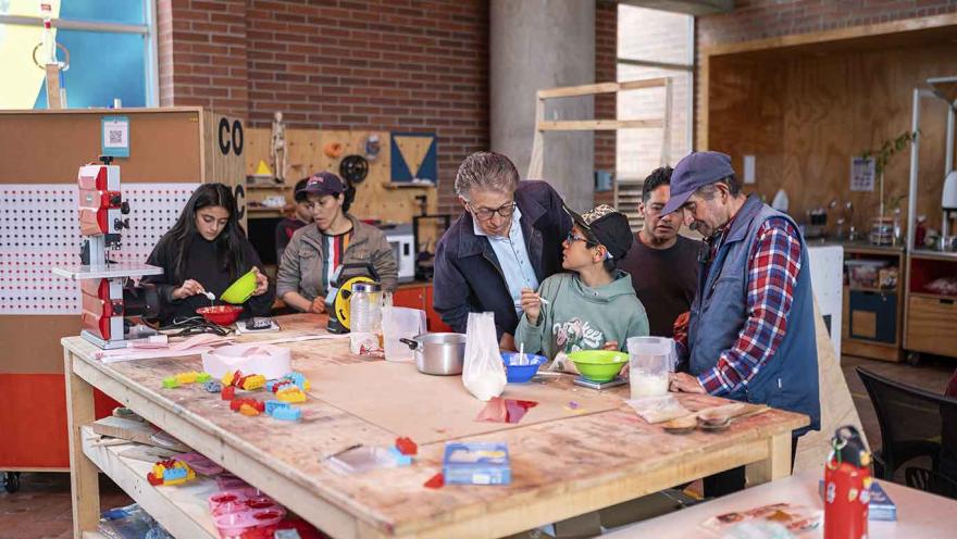Grupo de personas de distintas edades participando en un taller en un espacio tipo laboratorio o biblioteca. Alrededor de una mesa de trabajo con materiales, utensilios y objetos coloridos, un adulto guía a un niño mientras conversan, y otros participantes mezclan ingredientes y realizan actividades manuales.