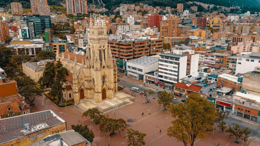 Vista aérea de una iglesia de estilo neogótico con torre alta y fachada ornamentada, ubicada en una amplia plaza urbana. Alrededor se observan árboles, peatones y edificaciones de diferentes alturas, con una densa zona urbana y montañas al fondo. La escena muestra un espacio público activo en la ciudad durante el día.