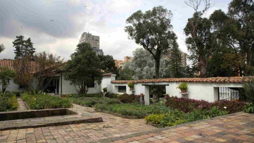 Jardín de una casa colonial con caminos de ladrillo, un pequeño estanque rectangular y muros blancos con tejas rojas, rodeado de árboles y vegetación, con edificios modernos visibles al fondo.