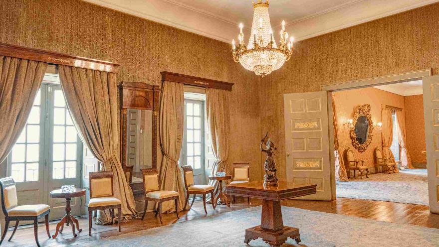  Interior de un salón elegante de estilo clásico, con paredes decoradas en tonos dorados y un candelabro de cristal colgando del techo. Hay muebles de madera y sillas tapizadas junto a ventanas con cortinas largas, y en el centro una mesa con una escultura. A través de una puerta abierta se observa otra sala con decoración similar y un gran espejo ornamental, creando un ambiente histórico y sofisticado.