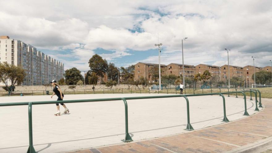 Pista de patinaje al aire libre rodeada por barandas verdes, donde una persona patina sobre el concreto. Al fondo se observan edificios residenciales, árboles y postes de luz bajo un cielo parcialmente nublado, en un entorno urbano amplio y abierto.