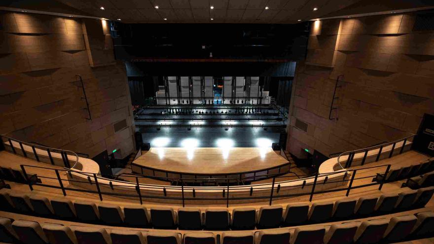 Vista desde las graderías del interior de un teatro moderno con asientos organizados en forma semicircular. Al frente se observa el escenario iluminado y la estructura técnica detrás del telón, con paredes en tonos cálidos y un techo con luces empotradas que iluminan el auditorio.