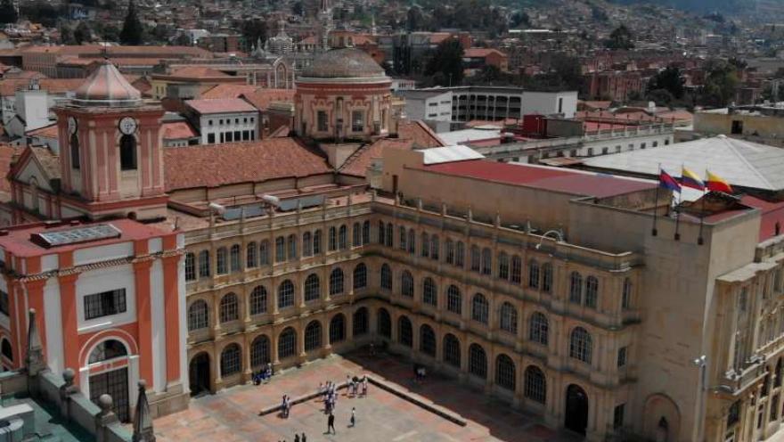  Vista aérea de un complejo histórico en el centro de la ciudad, con un amplio patio interior rodeado por un edificio de estilo colonial de varios niveles y arcos. A un lado se observa una iglesia con torre y cúpula, y en el patio hay pequeños grupos de personas.