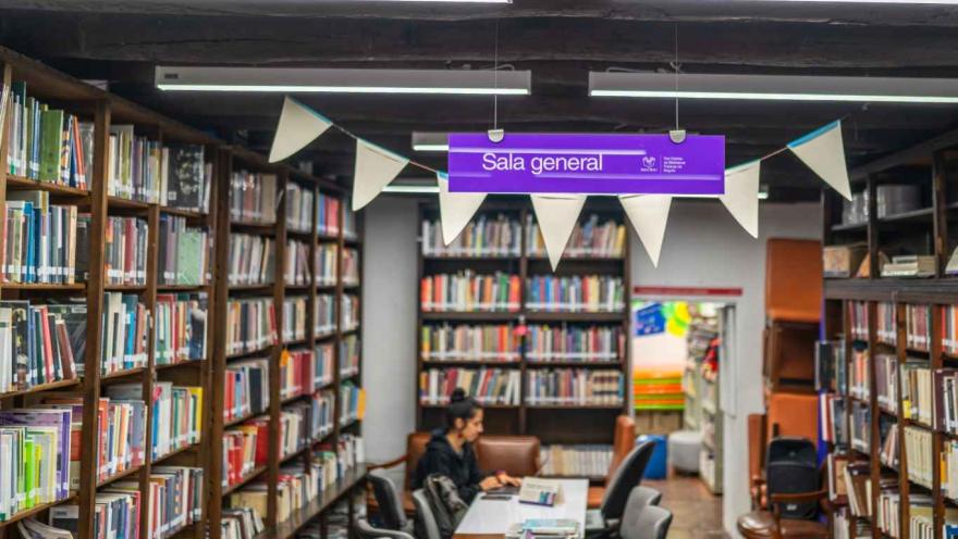  Interior de una biblioteca con estanterías de madera llenas de libros a ambos lados. En el centro hay una mesa larga con varias sillas donde una persona está sentada leyendo o trabajando con un libro. En la parte superior cuelga un letrero morado que dice “Sala general”, acompañado de banderines decorativos y luces fluorescentes en el techo. Al fondo se observan más estanterías y un espacio de lectura.