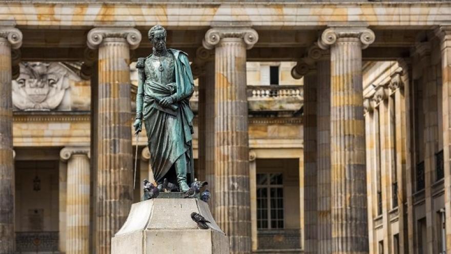 Estatua de un personaje histórico sobre un pedestal de piedra, vista de perfil, frente a un edificio de estilo colonial con fachada amarilla, ventanas simétricas y balcones decorados. En la base de la escultura hay algunas palomas, y al fondo se observan árboles que enmarcan este entorno urbano de carácter histórico.