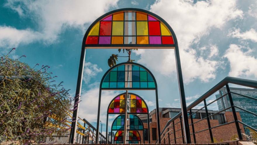 Estructura artística en forma de arcos metálicos con paneles de vidrio de colores, dispuesta en secuencia sobre un sendero. Los vitrales en tonos rojo, amarillo, azul y verde contrastan con el cielo parcialmente nublado al fondo. A los lados se observan barandas, vegetación y edificaciones urbanas, creando un espacio moderno y visualmente llamativo.