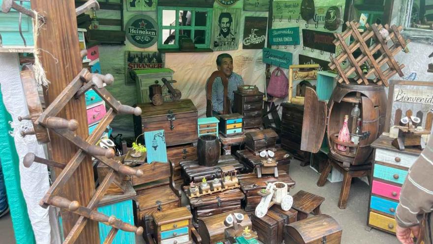 A craft market stall filled with decorative wooden objects, such as boxes, chests, small furniture, and carved figures. The pieces have rustic finishes and some details painted in vibrant colors.