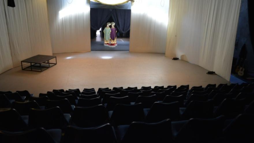 Interior de una sala de teatro con filas de sillas oscuras frente a un escenario sencillo cubierto con cortinas blancas. En el fondo del escenario, a través de una abertura con telones oscuros, se observan figuras o vestuarios coloridos dispuestos como parte de la escenografía.