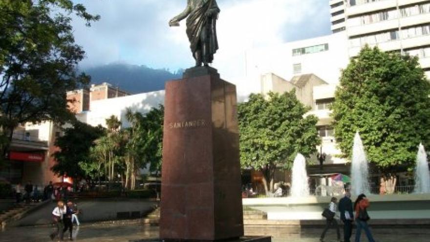 Estatua de bronce de un personaje histórico sobre un pedestal alto en una plaza urbana, con la inscripción “Santander” visible en la base. Alrededor se observan fuentes de agua en funcionamiento, árboles y edificios altos, mientras varias personas caminan por el espacio.