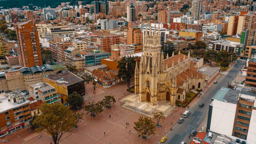 Vista aérea de una iglesia de estilo neogótico con torre central elevada y fachada detallada en piedra, ubicada en medio de una amplia plaza peatonal. Alrededor se observan árboles, personas caminando y vías con tráfico vehicular, además de numerosos edificios residenciales y comerciales que conforman un entorno urbano denso, con montañas visibles al fondo.