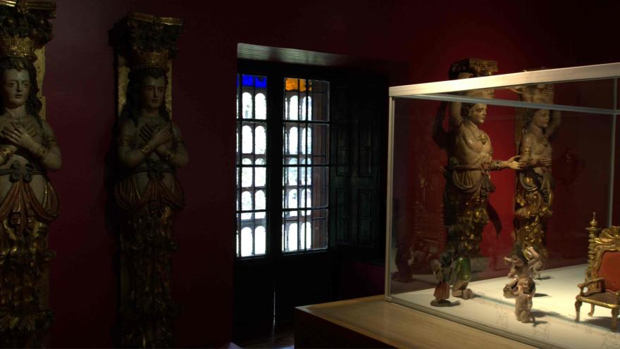 Sala de museo con paredes rojas donde se exhiben esculturas religiosas antiguas; a la izquierda hay figuras talladas de estilo barroco y, a la derecha, una vitrina de vidrio con pequeñas piezas.