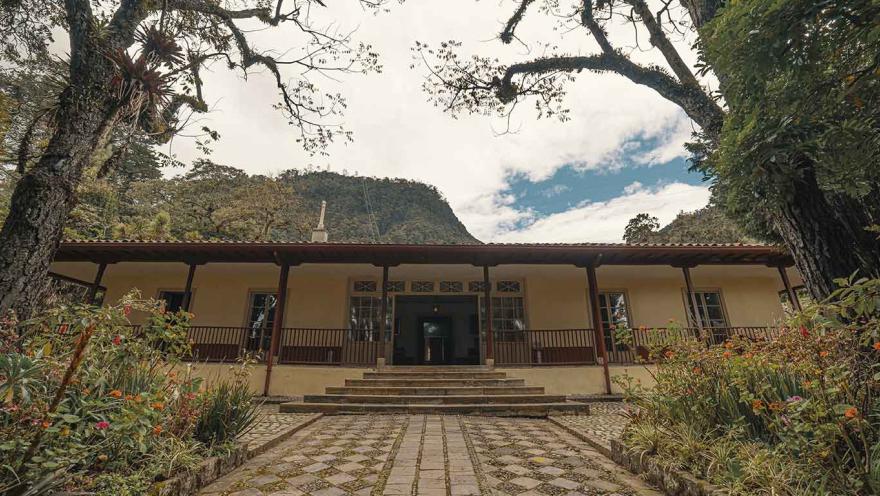 Vista frontal de la Quinta de Bolívar, una casa histórica de estilo colonial rodeada de jardines y altos árboles. Un camino empedrado conduce a la entrada principal con escaleras y un corredor con barandas de madera, mientras al fondo se observan las montañas verdes que rodean el lugar.