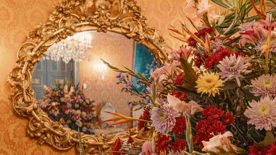 Primer plano de un arreglo floral colorido con flores rojas, rosadas, amarillas y naranjas, dispuesto en un elegante salón de estilo clásico. Al fondo se observa un gran espejo con marco dorado ornamentado que refleja un candelabro de cristal y más flores, creando un ambiente sofisticado y decorativo.