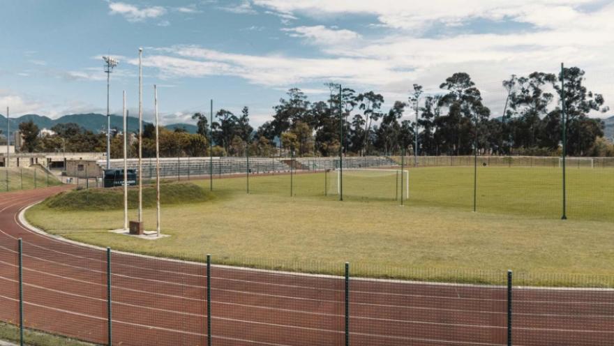 Escenario deportivo al aire libre con pista atlética roja que rodea una cancha de fútbol de césped verde, cercada con malla metálica. Al fondo se ven graderías, árboles altos y montañas bajo un cielo parcialmente nublado.